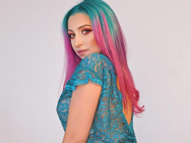 Agaa, con un cabello de color rosa a verde, vistiendo una blusa de encaje color turquesa, mirando por encima del hombro, mostrando sus pechos a través del encaje, con sombra de ojos roja y un piercing en la nariz..