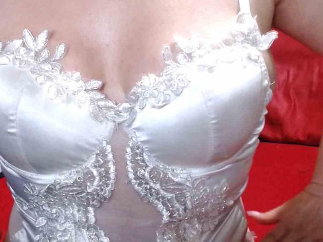 La lingerie en dentelle blanche de PriscillaHotMature, mettant en valeur ses seins volumineux et brillants avec des motifs floraux complexes et une partie centrale transparente, posée sur une surface rouge..