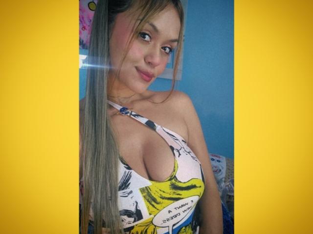 Melissa Arango, con lunghi capelli grigiastri, sorride, indossando un top bianco con spalline che lascia scoperte le spalle, con scritte a tema cartone animato in giallo e nero, mostrando il suo seno abbondante. Bordo giallo attorno all'immagine..