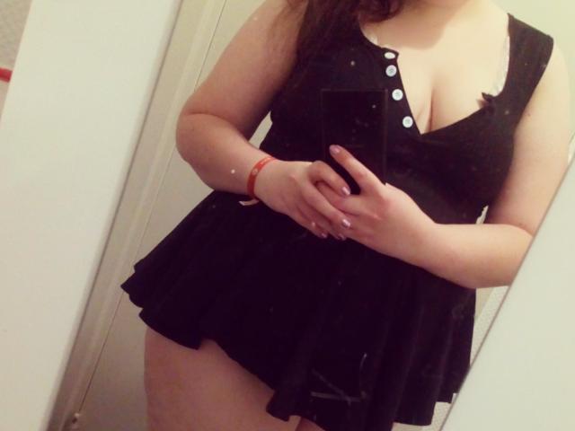 LoveClo trägt ein schwarzes Kleid mit Knöpfen, hält ein Telefon, zeigt ihre Brüste, trägt ein rotes Armband, Spiegel-Selfie, Haut ist sichtbar, steht mit verschränkten Armen, braunes Haar..