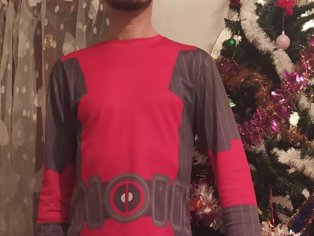 PierceBrick draagt een rood en grijs kostuum van een Marvel-film met een rond embleem, en staat voor een versierde kerstboom. Borsten niet zichtbaar, gezicht buiten beeld..