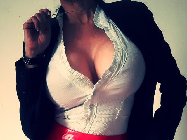 Uma mulher chamada LaParisienne, com seios grandes, vestindo uma camisa branca com babados e botões, um casaco preto e calças vermelhas, segurando um cigarro e usando uma pulseira preta..
