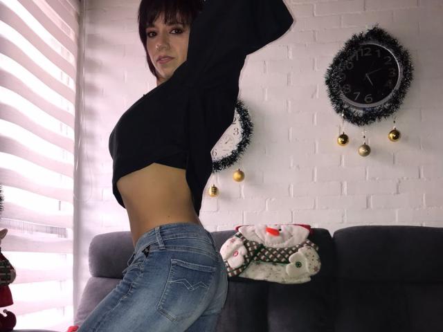 BellaLolita con cabello corto castaño, una blusa negra que deja ver su abdomen, jeans azules, de pie de lado, mostrando su trasero, en una habitación con un reloj y adornos..