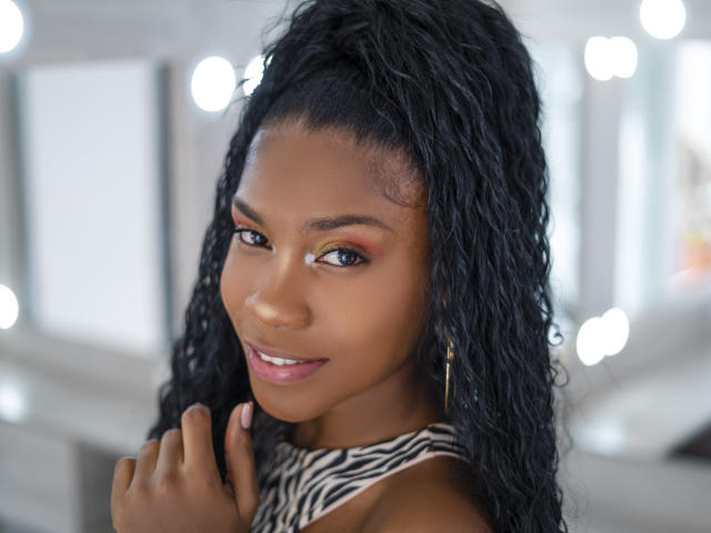 AlanaPatel has dark skin, long black curly hair in a high ponytail, and wears a zebra-patterned top, with a subtle smile and pink lipstick. She's touching her chin.