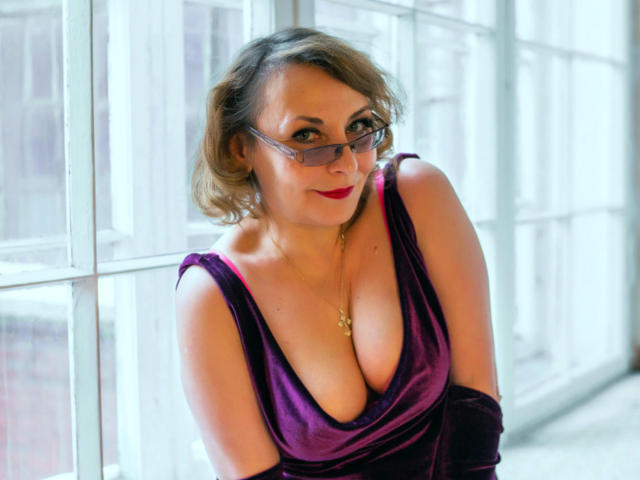 LadyIren mit Brille, lila Samtkleid, großer Brust, rotem Lippenstift, kurzen blonden Haaren, goldener Halskette, lächelnd, stehend am Fenster, weiches, natürliches Licht, sexy, selbstbewusst, süß..