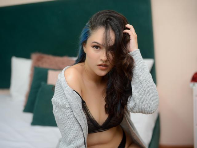 Stephanie Valdez, con riflessi blu nei suoi capelli scuri, indossa un cardigan grigio portato a spalla, un reggiseno di pizzo nero, e mostra il seno, guardando seducentemente la fotocamera..