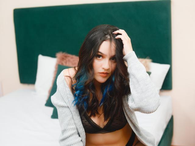 Stephanie Valdez, met blauw getipt haar, draagt een grijze cardigan en een zwarte kanten BH, met haar hand in haar haar, een verleidelijke blik, op een bed met een groene hoofdeinde en witte kussens..