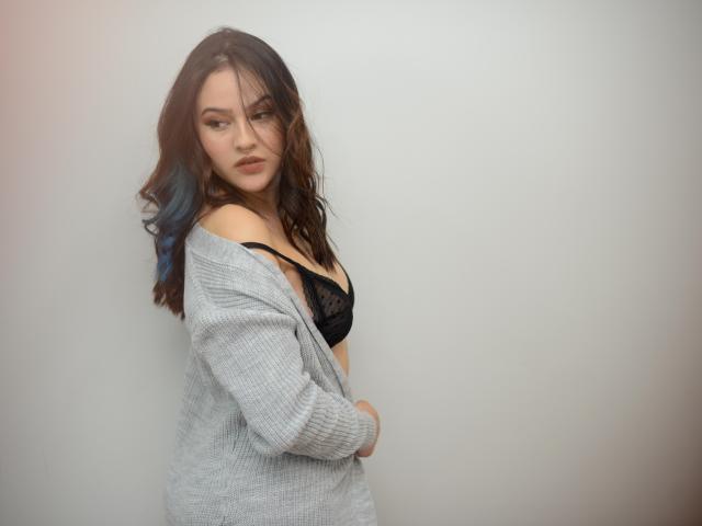 Stephanie Valdez mit blau gefärbtem Haar, einem grauen Pullover, einem schwarzen Spitzen-BH, der die Brüste zeigt, mit verschränkten Armen, ernster Miene, welligem braunem Haar, heller Haut und einem weißen Hintergrund..