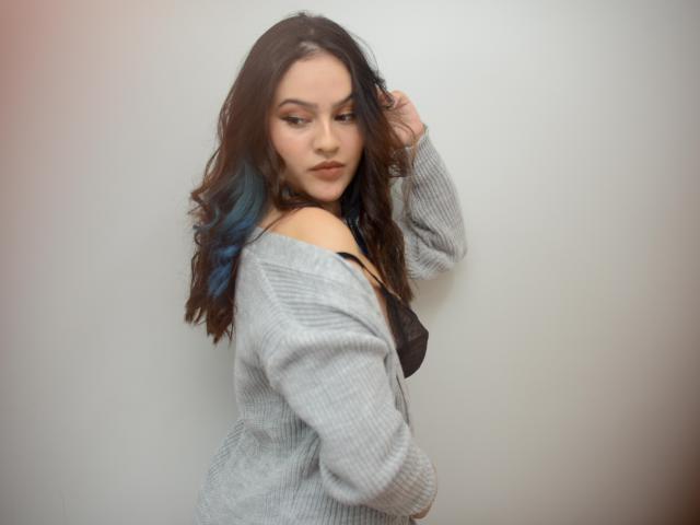 Stephanie Valdez, con cabello castaño ondulado y reflejos azules, vestida con un suéter gris, un sostén negro, y mirando por encima del hombro con una ligera sonrisa, mostrando su trasero..