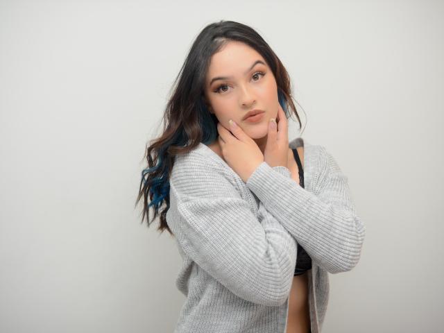 Stephanie Valdez, con cabello de punta azul, vistiendo un cárdigan gris de punto, un sujetador negro, con las manos cerca del rostro, expresión neutra, piel clara, ojos marrones, maquillaje suave, cabello ondulado, ligera sonrisa..