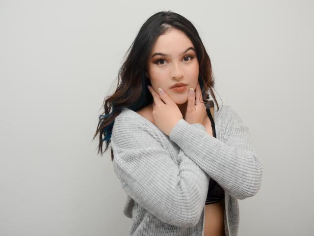 StephanieValdez with wavy dark brown hair, gray ribbed sweater, hands near face, brown eyes, subtle makeup, light skin, black bra visible, looking at camera, neutral expression.
