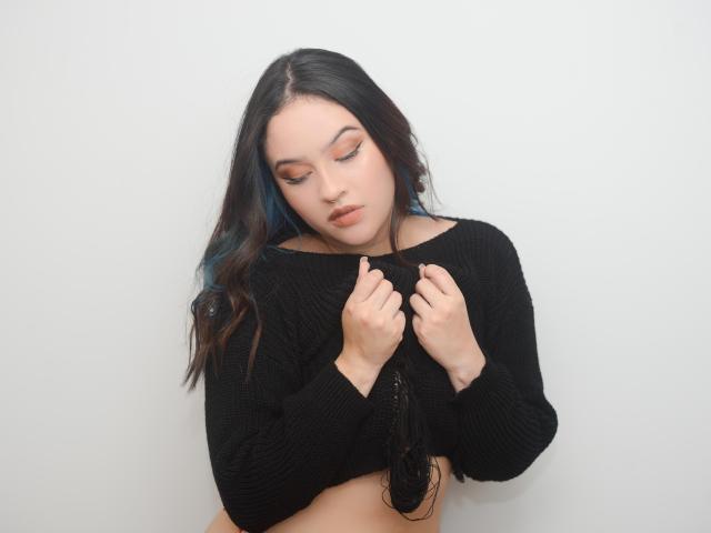 Stephanie Valdez, con cabello largo y oscuro, vestida con una blusa de punto negra, con las manos en el pecho, ojos cerrados, mirando hacia abajo, mostrando sus senos, en una foto suave con iluminación blanca..