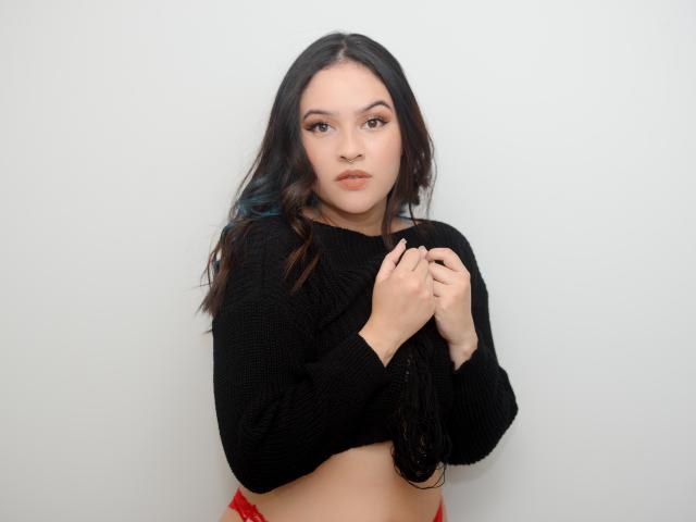 Stephanie Valdez, con senos de tamaño medio, vistiendo un suéter negro, con las manos en el pecho, ropa interior roja, piel clara, ojos marrones, cabello oscuro ondulado, reflejos azules sutiles, expresión neutra, fondo blanco..