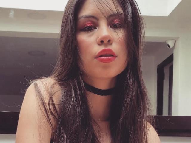 IsaTimeSweet with long brown hair, red lipstick, and black choker, looking at the camera with a slight pout, wearing a black top, in a low-light indoor setting.