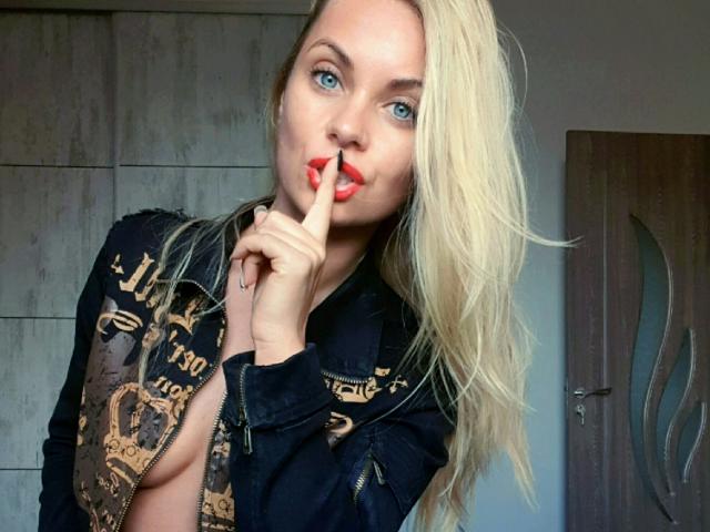 Blonde AlisaRose with blue eyes, red lips, and slightly open black jacket, showing finger to mouth, exposing one tit, wearing a dark jacket with gold embroidery.