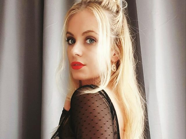 Blond AlisaRose with red lipstick, wearing a sheer black top, looking over her shoulder, blue eyes, and long hair in a ponytail. Her tits are visible through the top.