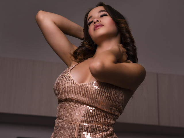 LuciRaven, con le braccia alzate, indossa un abito rosa scintillante ricoperto di paillettes, che mette in mostra il suo seno grande e formoso, e ha un'espressione sicura e sensuale, con capelli castani ondulati e rossetto rosa..