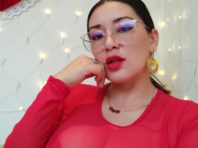 Maloryxx, con lápiz labial rojo, gafas y aretes dorados, vestida con una blusa roja transparente, tiene una mano cerca de su boca, mostrando sus senos y un collar..