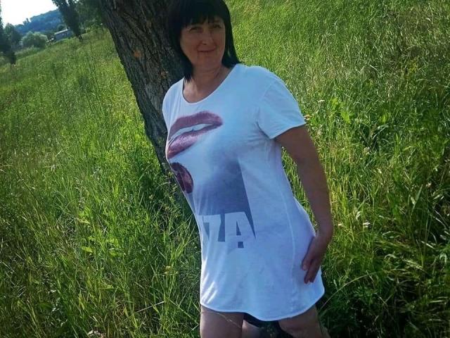 NikaMel mit schwarzen Haaren, die ein weißes T-Shirt trägt, auf dem rosa Lippen und ein Buchstabe "A" abgebildet sind, steht auf grünem Gras. Sie trägt schwarze Unterwäsche, die sichtbar ist, und hat einen leicht rundlichen Hintern..