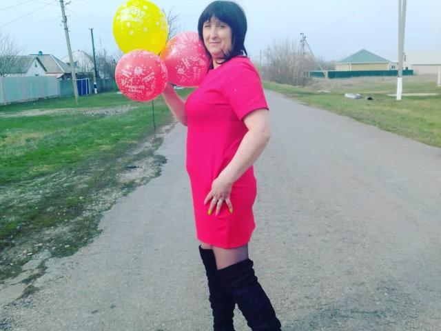 NikaMel se tient sur un chemin de gravier, vêtue d'une robe rose, de bottes noires, et tenant des ballons rouges et jaunes, avec des cheveux noirs courts et un léger sourire..