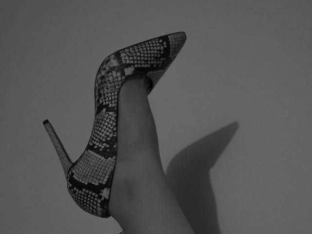 La jambe d'AbigailCroixX, dans une chaussure à talon aiguille noire et blanche à motifs, avec son pied pointé vers le haut et une ombre projetée sur le mur. Son talon est pointu et elle porte des escarpins..