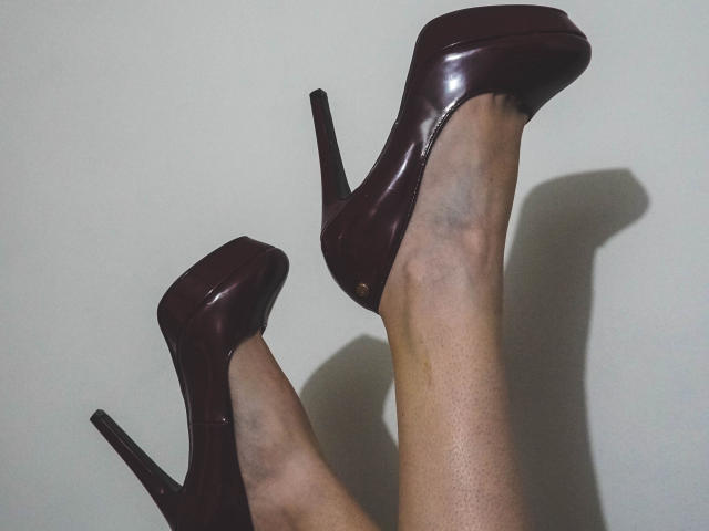 Las piernas de AbigailCroixX, con tacones altos de color marrón oscuro, con los pies cruzados a la altura de los tobillos, mostrando sus tacones de aguja brillantes y relucientes, y su piel ligeramente azulada..