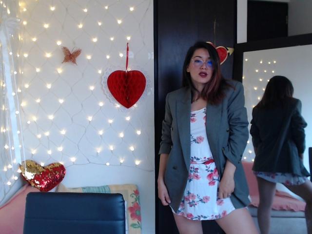 Maloryxx, avec des cheveux teints en rouge et portant une robe à fleurs, un blazer vert, debout devant un miroir, montrant ses fesses, entourée de guirlandes lumineuses, d'un cœur rouge et de décorations murales en forme de papillons..