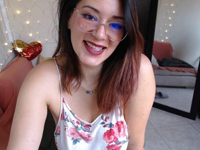Maloryxx heeft een bril met een blauwe rechterlens, ze glimlacht met donkerrode lippenstift, draagt een bloemenprint topje en heeft bruin haar dat tot op haar schouders reikt. Haar borsten zijn klein en ze is schattig..