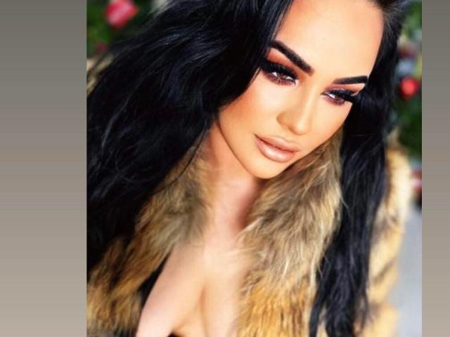 HottyNicole with long black hair, heavy eyeliner, and nude lipstick, wearing a fur coat that's open to show her tits. Her eyes are half-closed, giving a sultry look.