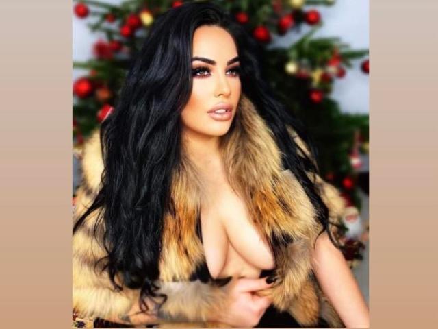 HottyNicole, con cabello negro largo, vistiendo un chaleco de piel, mostrando sus grandes senos, de pie frente a un árbol de Navidad con adornos rojos, luciendo sexy con ojos seductores..