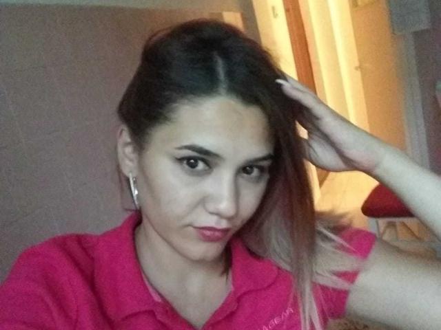 LovelyFace con capelli castano scuro, una camicia rosa e un trucco leggero, che si tiene i capelli, guarda direttamente nella fotocamera, mostrando il suo viso carino e le labbra rosse..