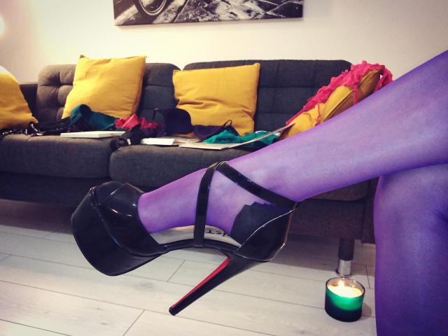 WandaDominatrix's purple pantyhose legs with black high heels, red soles, crossed on a grey sofa. Yellow pillows, green bag, candle on white floor in background.