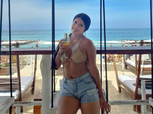 Valentina Gomez met een bruine bikini top die haar borsten laat zien, draagt denim shorts, houdt een cocktail met een banaan vast, glimlacht, staat op een balkon aan het strand, heeft lang zwart haar, de lucht is blauw..