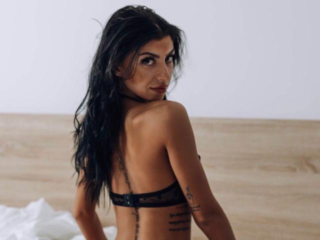 KateSparks, con lunghi capelli neri, che si guarda indietro, indossa un reggiseno di pizzo nero, mostrando tatuaggi sulla schiena e sul braccio, con i seni e i glutei leggermente visibili..