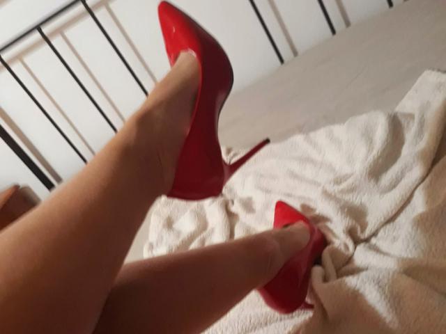 Aiyana's legs with shiny red high heels, feet pointed upwards, on a white textured blanket, black metal bed frame in background. Her smooth, tan skin contrasts with the red heels.