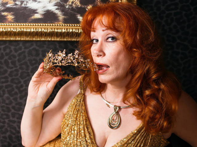 Une femme séduisante aux cheveux roux bouclés, portant une robe dorée pailletée et un collier, léchant une couronne florale dorée et rose, montrant ses seins, dans une pose sexy et aguicheuse, avec une expression espiègle..