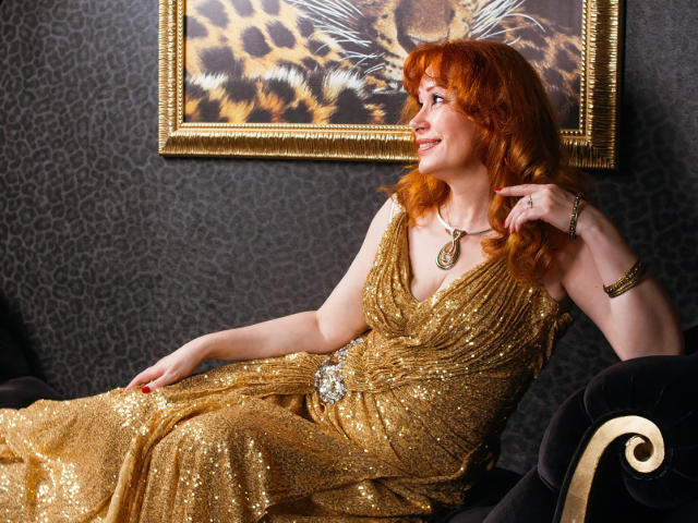Mujer atractiva con cabello rojo, vestido dorado brillante y collar, sentada en un sofá de terciopelo negro, mirando hacia un lado, sonriendo, con la mano en la barbilla, mostrando ligeramente su trasero..