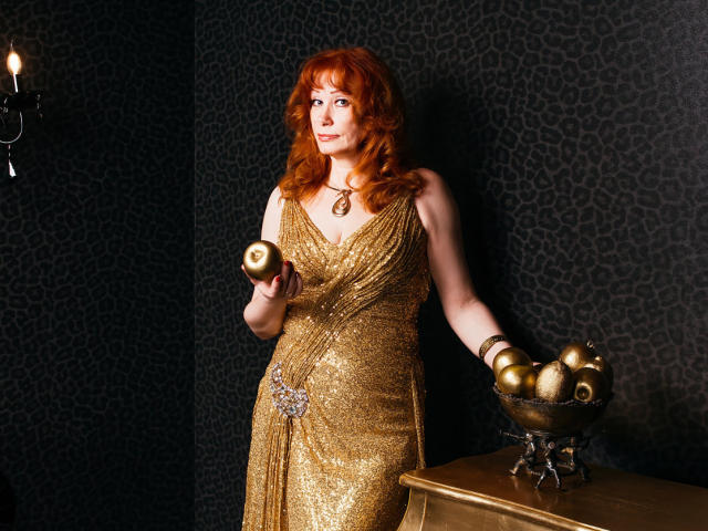 Eine Frau mit roten Locken, die ein glänzendes goldfarbenes Kleid trägt, hält einen goldenen Apfel in einer Hand und eine Schüssel mit goldenen Äpfeln in der anderen, und steht in der Nähe eines Holztisches..