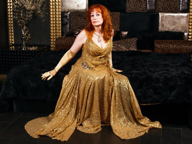 Een aantrekkelijke vrouw met rood haar, gekleed in een glinsterende gouden jurk, zittend op een zwart bed, met gele handschoenen, die haar borsten en billen laat zien, met een sexy en zelfverzekerde blik..