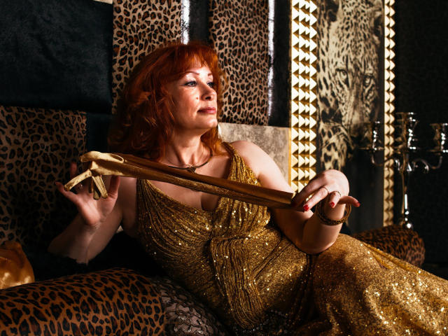 Una donna con i capelli rossi, vestita con un abito dorato scintillante, che tiene un nastro dorato, seduta su una sedia con motivo leopardato, che guarda verso l'alto con un leggero sorriso..