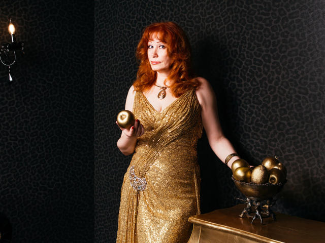 Mujer de cabello rojo, con un vestido dorado y brillante, sosteniendo una vela, de pie junto a una mesa con un candelabro de latón, con los senos ligeramente visibles, mirando hacia un lado..