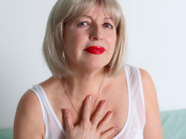 SamanthaLala with blonde bob, red lipstick, white tank top, gold necklace, and hand on chest, looking slightly up. Boobs partially visible. Soft lighting, slight smile.
