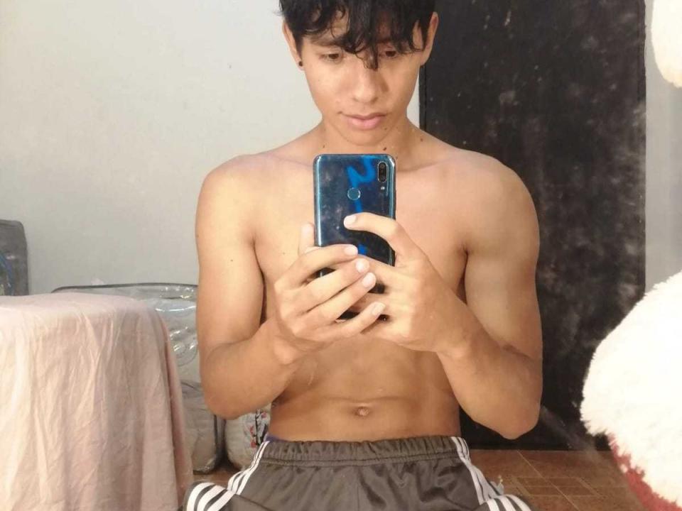 AndrewGregory, zonder shirt, met een getraind, gespierd lichaam, houdt een blauwe telefoon vast en maakt een spiegelzelfportret, draagt grijze korte broek, kijkt naar beneden naar de telefoon, kort zwart haar, bruine huid..
