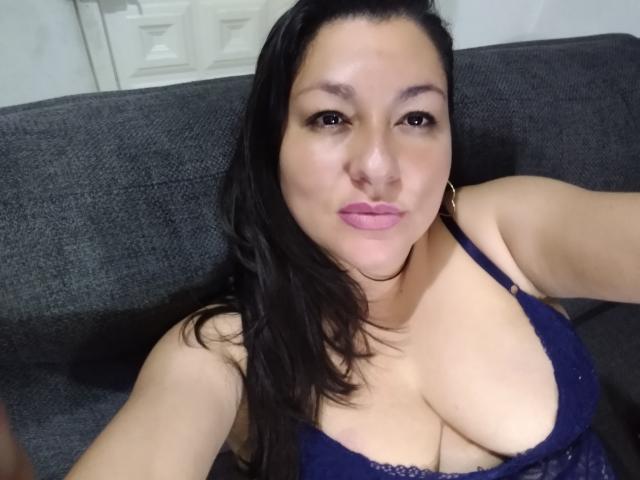 SakotoDika with dark hair, wearing a blue lace bra, showing her large boobs, pouty pink lips, and hoop earrings, sitting on a gray sofa.