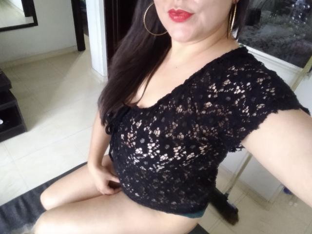 SakotoDika with long dark hair, red lipstick, gold hoop earrings, black lace top, showing her tits, sitting with her hand on her green panties, in a selfie.