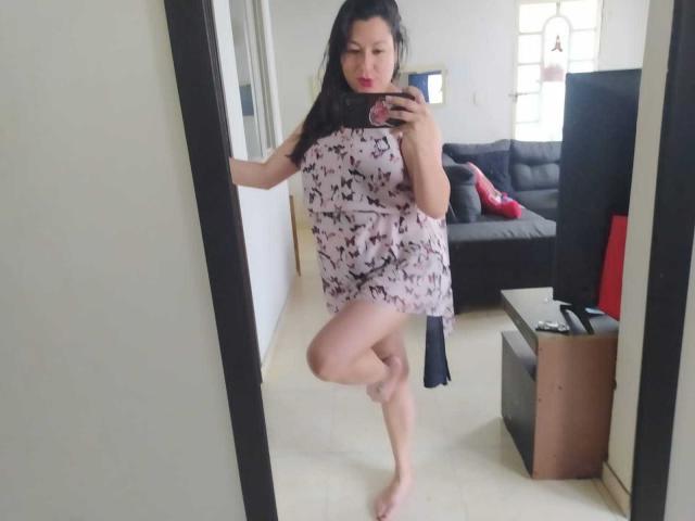 SakotoDika, barefoot, wearing a pink and black butterfly-patterned dress, standing on one leg, taking a mirror selfie, black hair, red lipstick, white room, black couch in background.