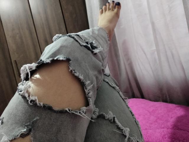 LuluFrancaise's ripped grey jeans show a large tear exposing her bare, pale-skinned butt. She has dark purple toenails and is resting her foot on a pink blanket.