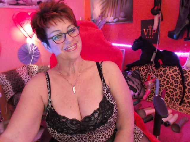Bettina with short brown hair, glasses, and a leopard print dress showing her big boobs, sitting in a red chair, smiling. Pink and red lights are on.
