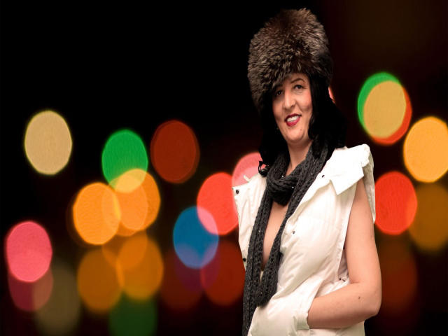 Ridemeraf with a fluffy brown fur hat, white puffy vest, black scarf, and red lipstick, smiling, hands in vest, colorful bokeh lights in the background.