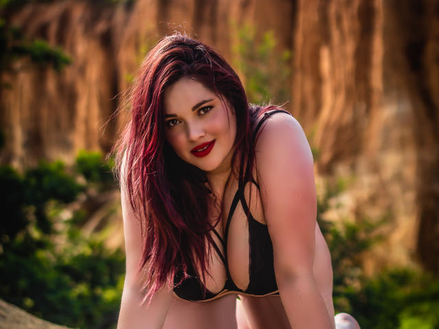 LissaClaire with red lipstick and black bikini, leaning forward, showing her big tits, and smiling at the camera with her purple hair falling over her shoulder.